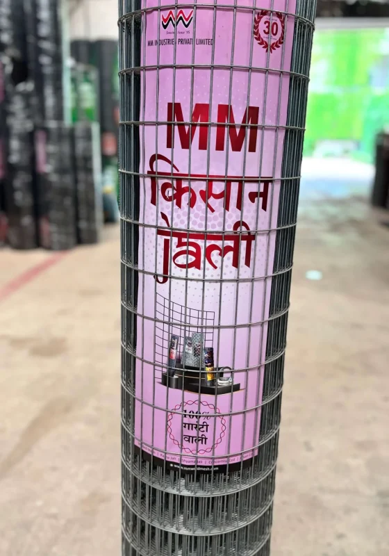 A roll of metal wire fencing stands upright, wrapped with a pink label that reads MM किसान जाली in Hindi, featuring an image of a tractor carrying fencing in the lower section of the label. This sturdy anti climb weldmesh design offers enhanced security and durability for agricultural and commercial applications.