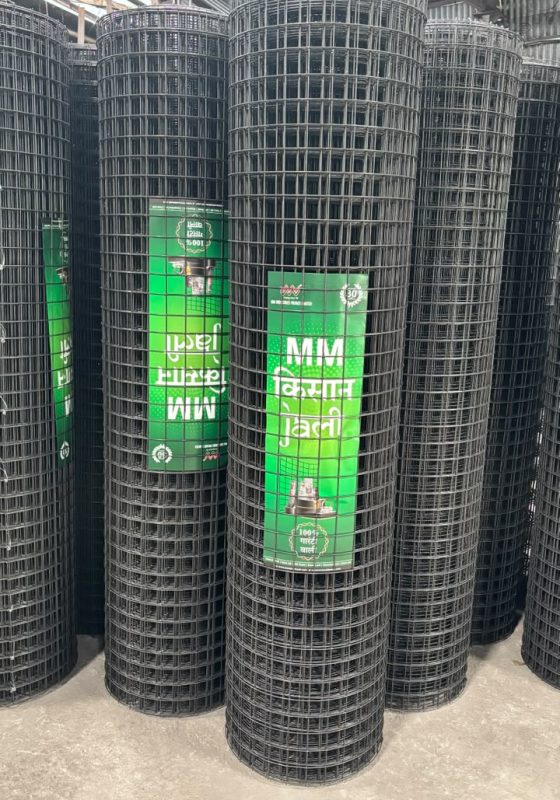 Black MS welded wire mesh rolls stacked vertically inside a warehouse, featuring MM किसान जाली branding. High-strength mild steel weld mesh designed for agricultural fencing, farm boundaries, industrial enclosures, construction reinforcement, and perimeter security.